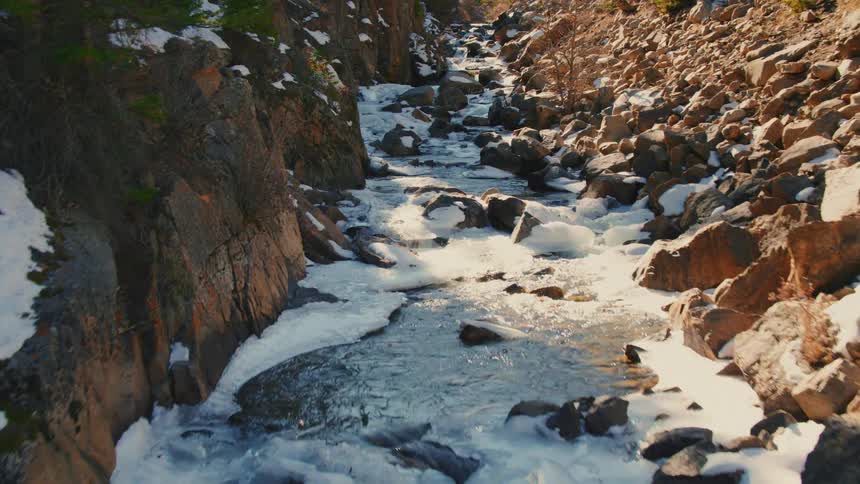 Drone footage slowly flies over a partially frozen mountain creek winding through a rocky canyon in the winter sun