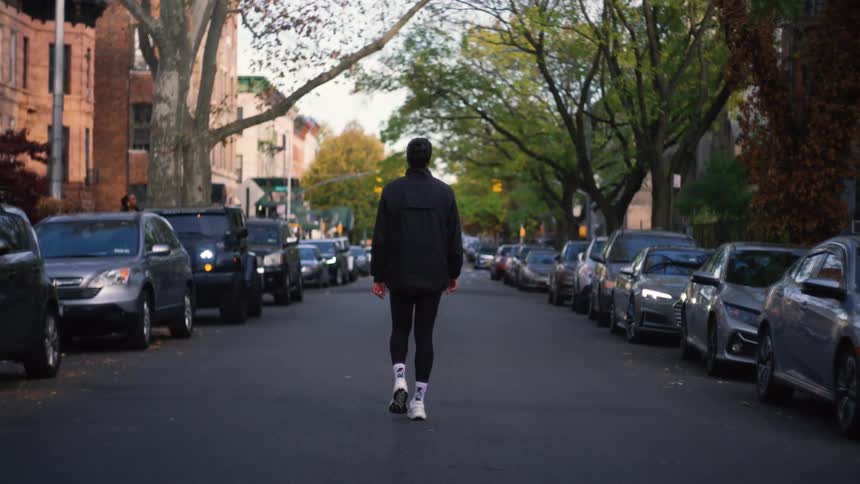 Slow motion rear view of a person in athletic gear walking down the middle of a quiet urban street lined with parked cars