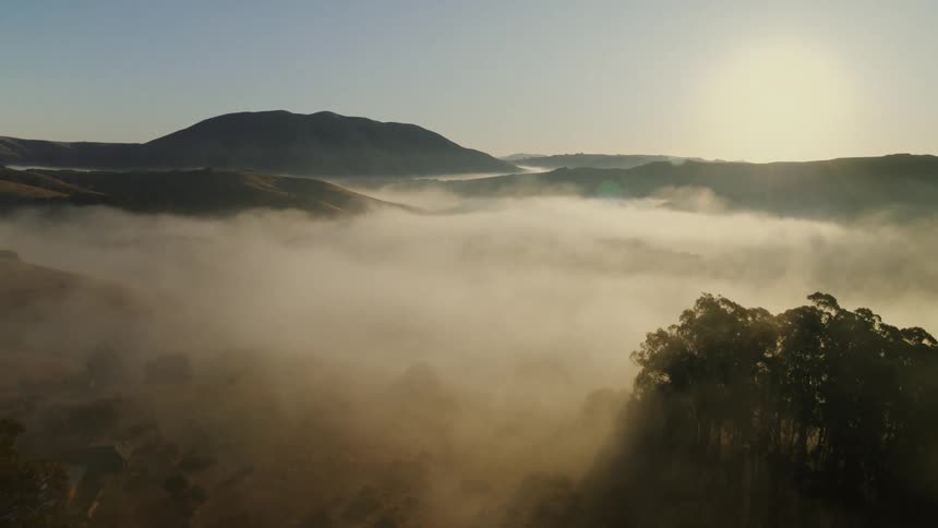 A slow pull-back aerial shot reveals a majestic mountain valley filled with dense fog during a golden sunrise