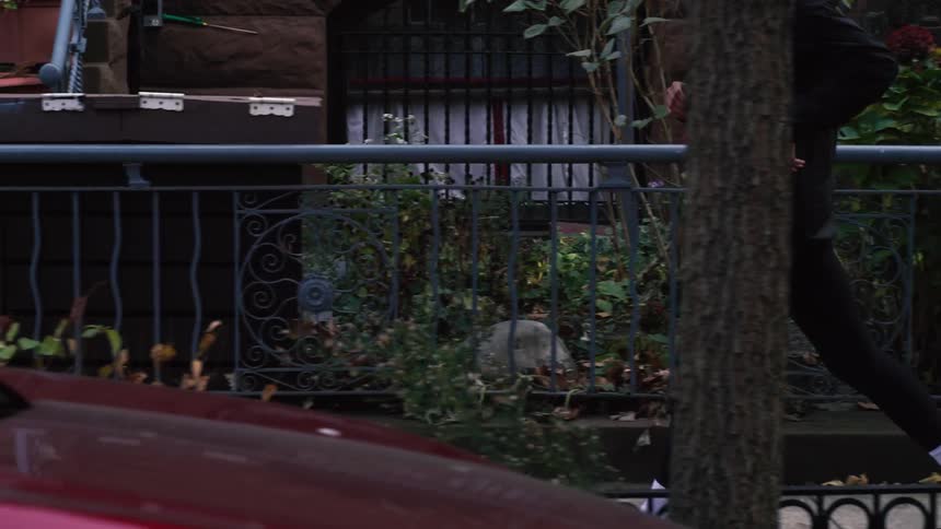A woman jogs in slow motion down a city street lined with cars and brownstone buildings