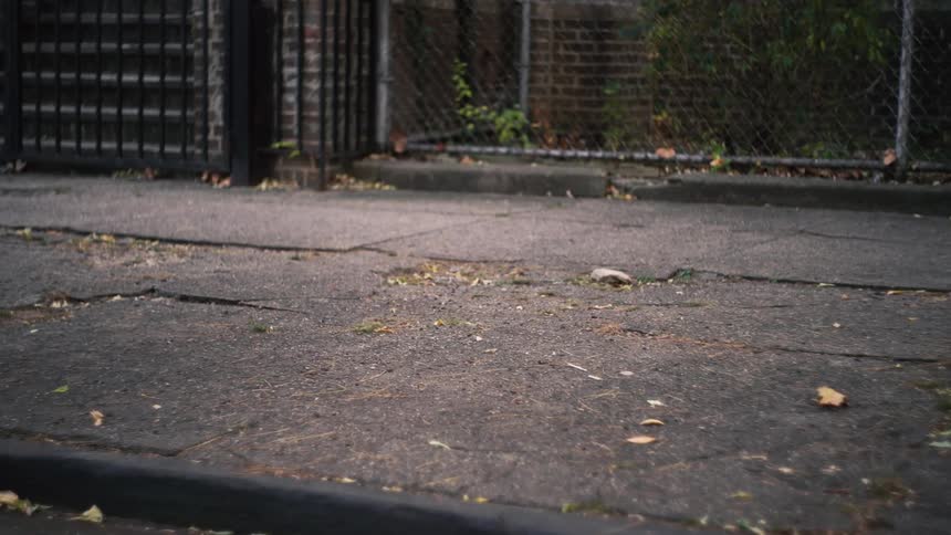 A low-angle, slow-motion shot of a person's feet in white sneakers walking on a cracked urban sidewalk
