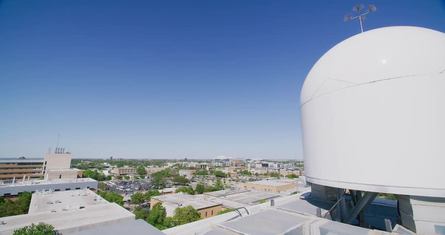A rooftop view of a city with a large white weather radar dome and anemometer under a clear blue sky