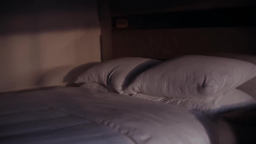 A close-up shot of a made bed with white pillows and a comforter gently rocking in a dimly lit room