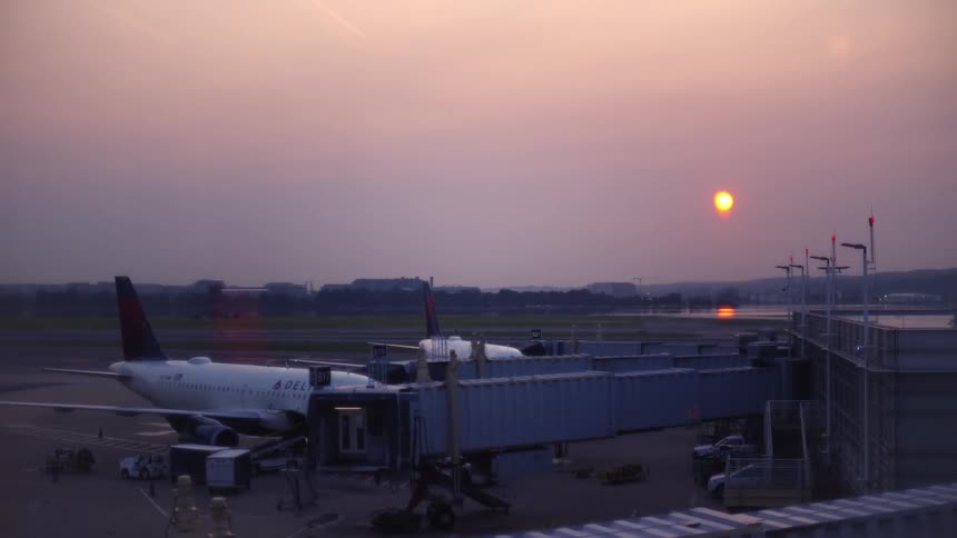 An airplane sits at the gate during a hazy sunset while another jet taxis on the runway in the background