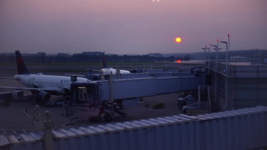 A passenger plane sits at the airport terminal gate as another plane takes off in the background during a beautiful hazy sunrise