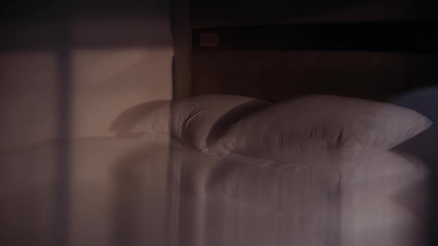 A moody close-up of an empty, made bed with white linens in a dimly lit room as a transparent curtain sways in the foreground