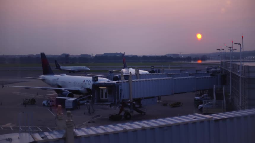 An airplane sits at the airport gate on the tarmac as another plane taxis in the background during a hazy sunset