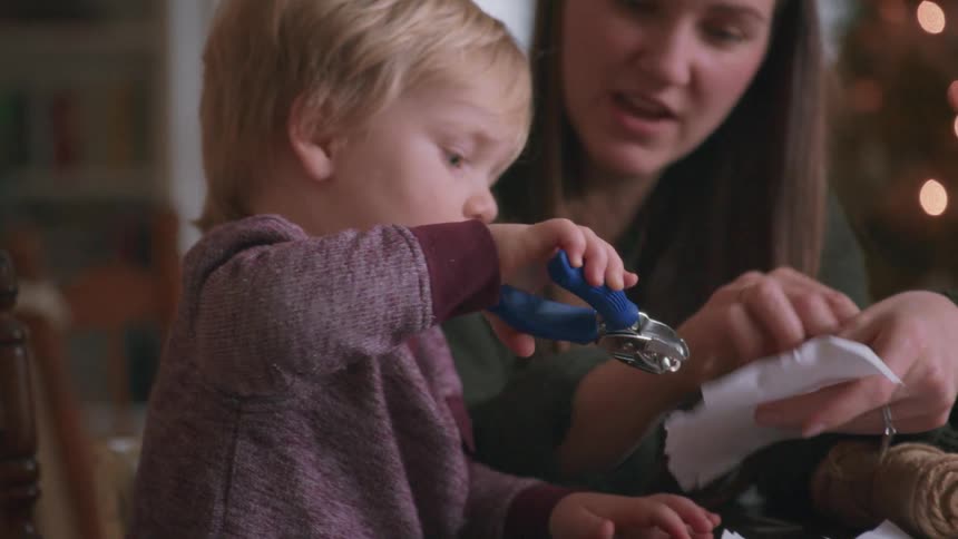 A sweet toddler carefully uses a hole punch with his mother's guidance to make homemade holiday crafts by the Christmas tree