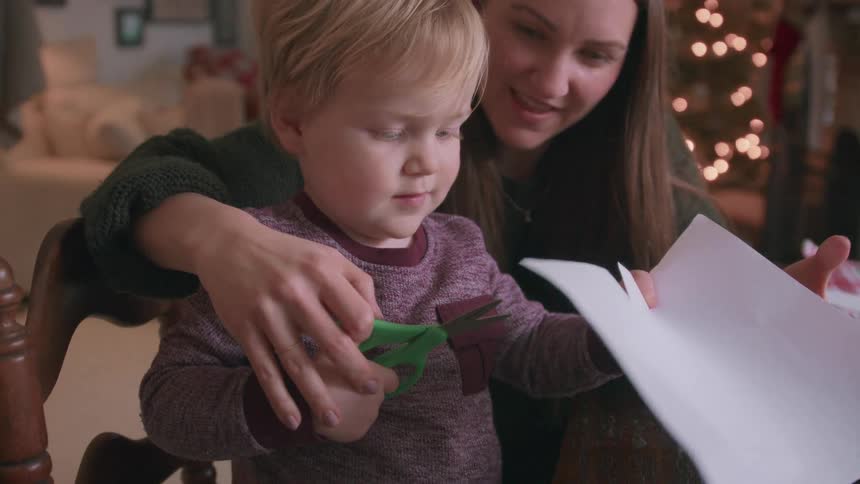 A mother helps her toddler son cut paper with scissors for a holiday craft in their cozy home