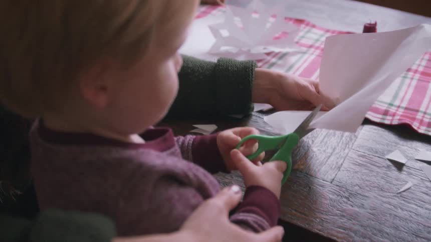 A mother helps her toddler learn to use scissors while making paper snowflakes together at a table for the holidays
