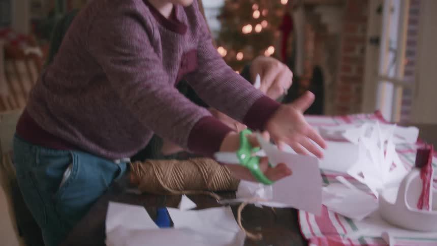 A focused toddler carefully cuts paper with safety scissors as his loving mother looks on while making Christmas decorations