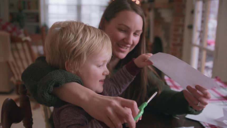 A loving mother smiles as she helps her young blonde-haired child learn how to cut paper with scissors to make Christmas snowflakes at a wooden table