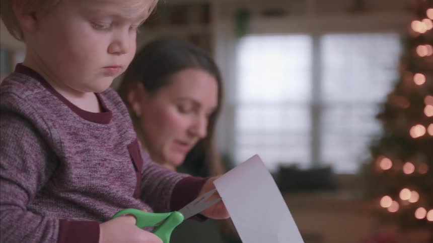 A cute young boy carefully cuts paper with green safety scissors as his mom looks on in front of a glowing Christmas tree