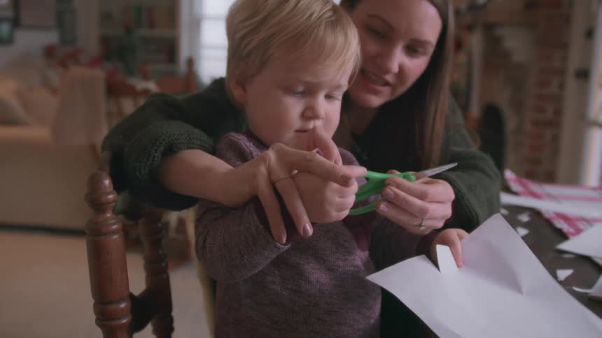 A young mother patiently teaches her toddler son how to cut paper with scissors in a cozy living room
