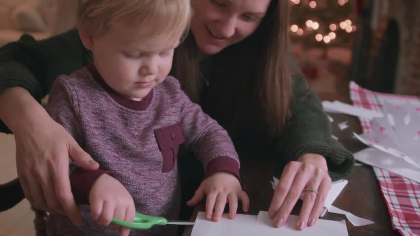 A mother and her young son cut paper snowflakes together at a table with a Christmas tree in the background