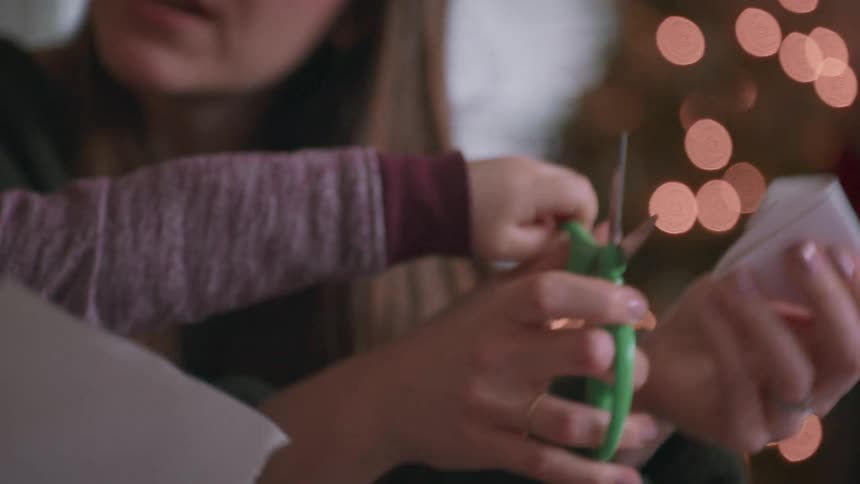 A mother patiently helps her young toddler son learn how to use scissors to make paper crafts in front of a Christmas tree