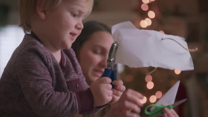 A cute toddler carefully makes Christmas crafts with his mom in slow motion with a beautifully lit tree in the background