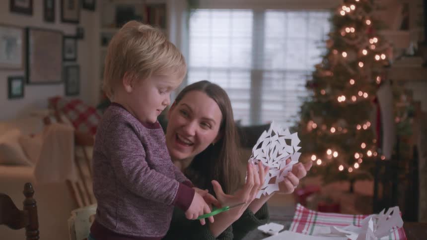 A smiling mother helps her young son cut out a paper snowflake in their festively decorated living room with a Christmas tree in the background