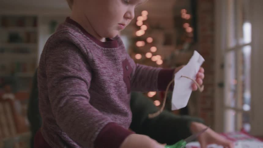 A focused toddler carefully uses safety scissors to cut paper for holiday crafts while his mom works in the background