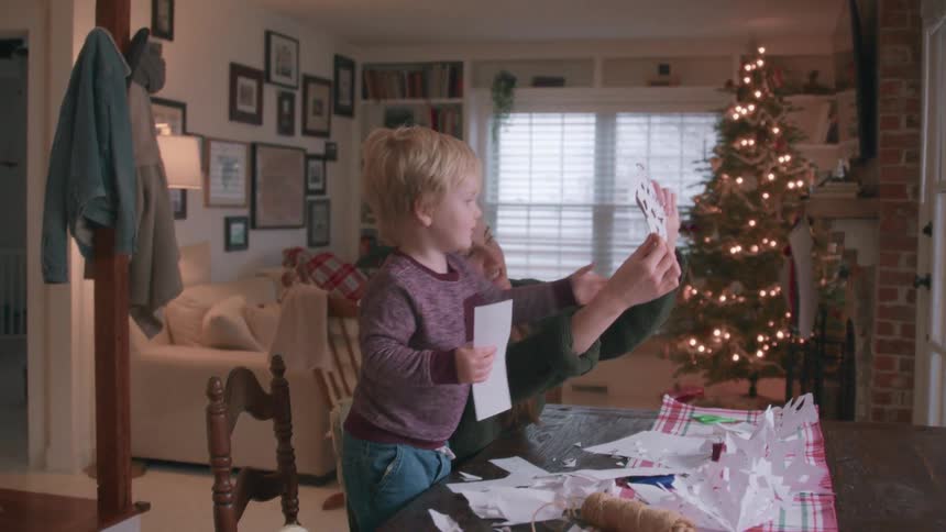 A mother and her young son enjoy making paper snowflakes together at the dining table with a lit Christmas tree in the background