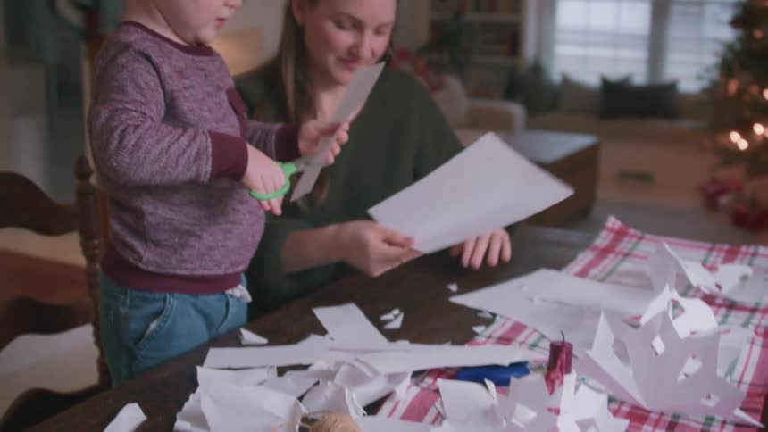 A young boy and his mother make paper snowflakes together at a dining table during the holidays