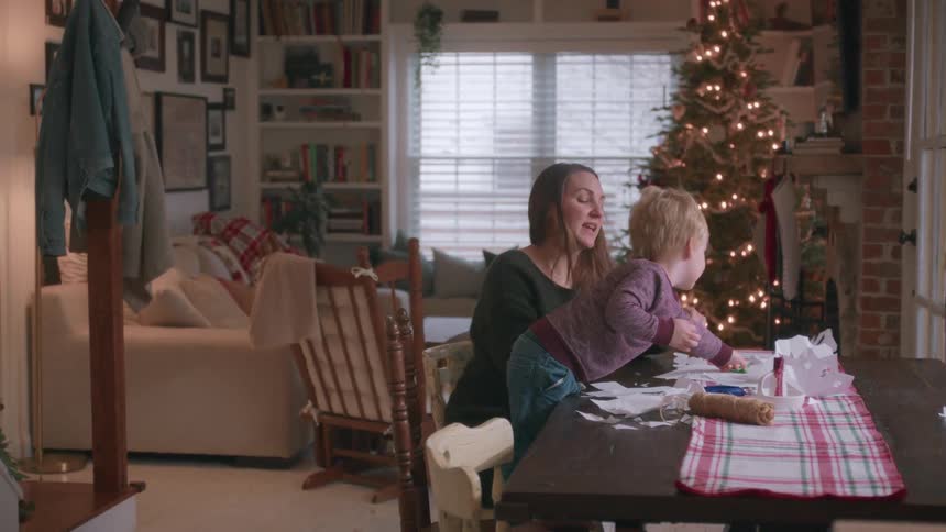 A young mother and her toddler son make paper snowflakes together at their dining room table with a Christmas tree in the background