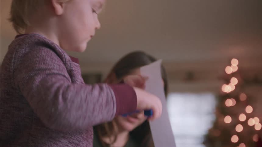 Cute blonde toddler concentrates on making a Christmas craft with a hole punch while his mother watches over him in their cozy home
