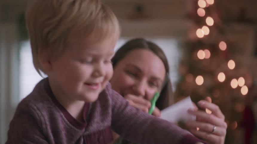 A mother helps her young son cut out paper snowflakes for Christmas decorations in slow motion with a glowing tree in the background