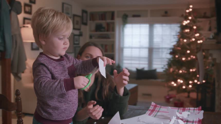 A mother patiently helps her adorable toddler son use scissors to cut paper snowflakes for Christmas decorations