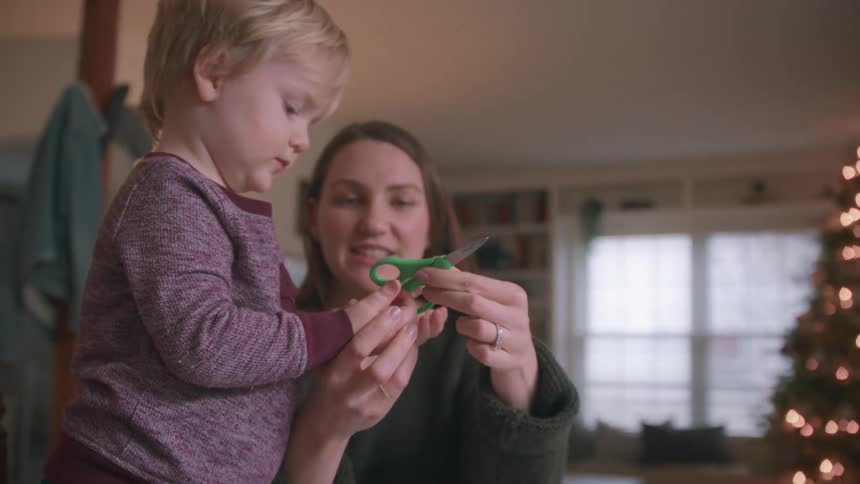 A mother patiently teaches her young toddler son how to use scissors for the first time in their cozy home