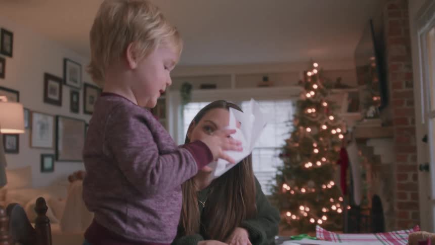 A mother and her cute toddler son make paper snowflakes together at a table in their cozy home with a Christmas tree in the background
