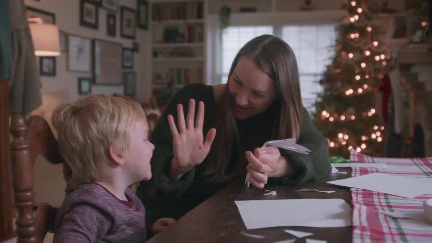 An excited young boy and his mother unfold a paper snowflake they made together, celebrating with a high-five in front of a Christmas tree