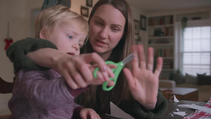 A loving mother teaches her toddler son how to use scissors to cut paper snowflakes for the holidays
