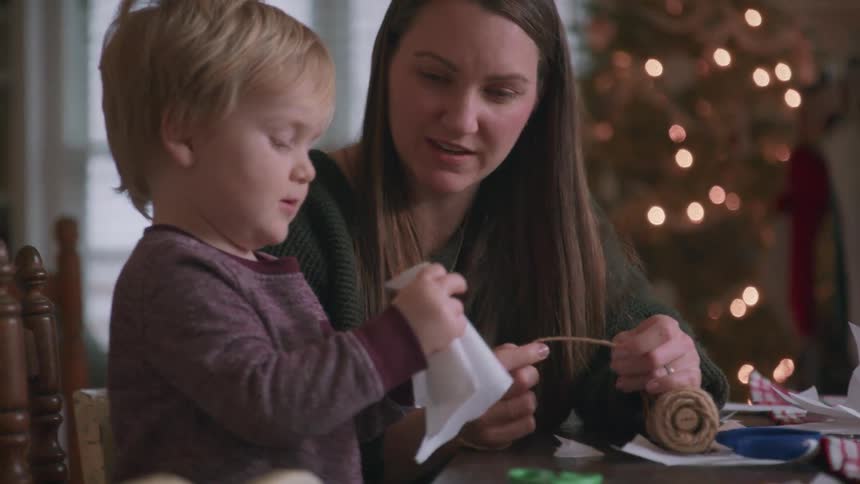 A mother and her young son enjoy a slow-motion moment making Christmas crafts together with a lit tree in the background