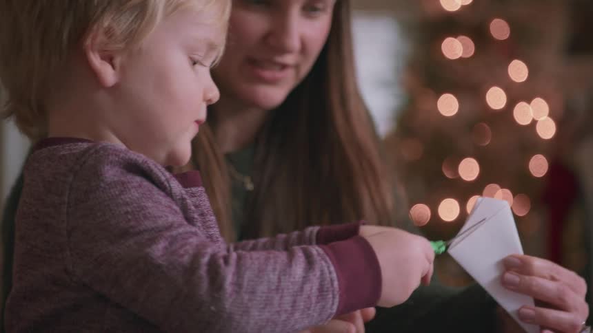 A mother patiently teaches her young son how to cut paper with scissors to make a Christmas snowflake decoration in slow motion