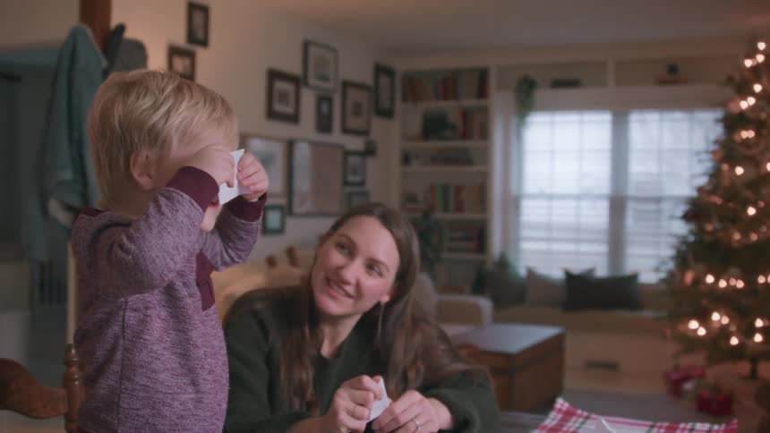 A mother smiles as her adorable toddler son helps her make paper snowflakes for Christmas, playfully covering his eyes with a paper scrap