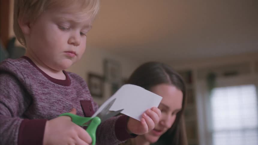 Close up of a young boy with blonde hair concentrating on cutting a piece of paper with green scissors indoors