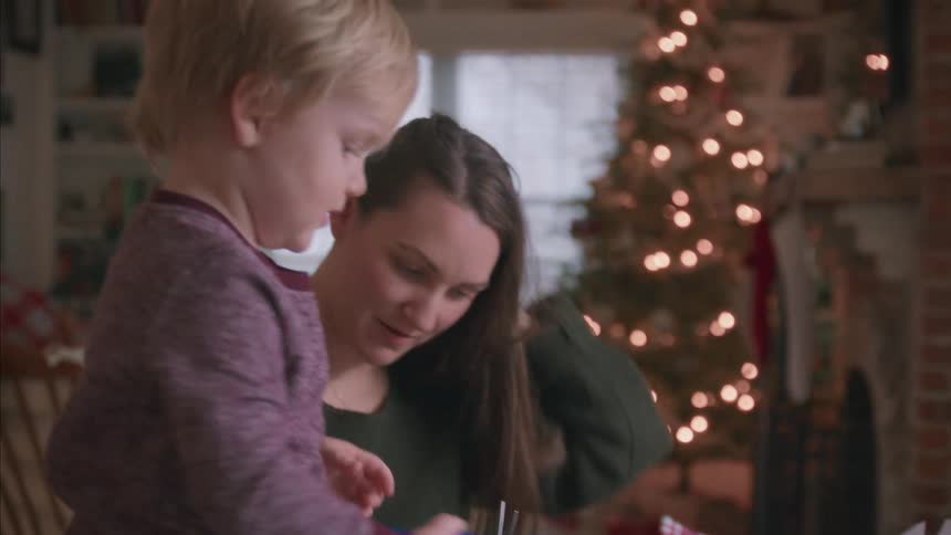 A young mother and her toddler son happily make Christmas crafts together in their cozy, festively decorated living room