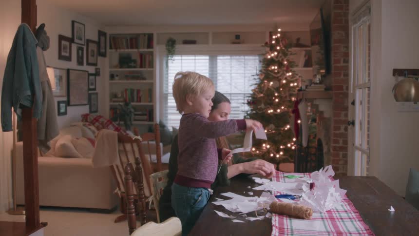 A young mother and her toddler son create paper snowflakes together at the dining table with a lit Christmas tree in the background