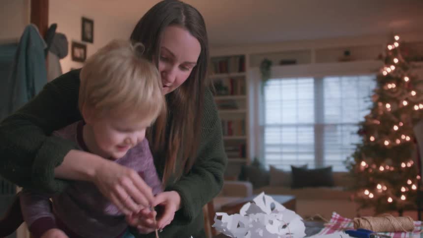 A joyful mother and her toddler son laugh while making paper snowflake decorations together in their cozy home by the Christmas tree