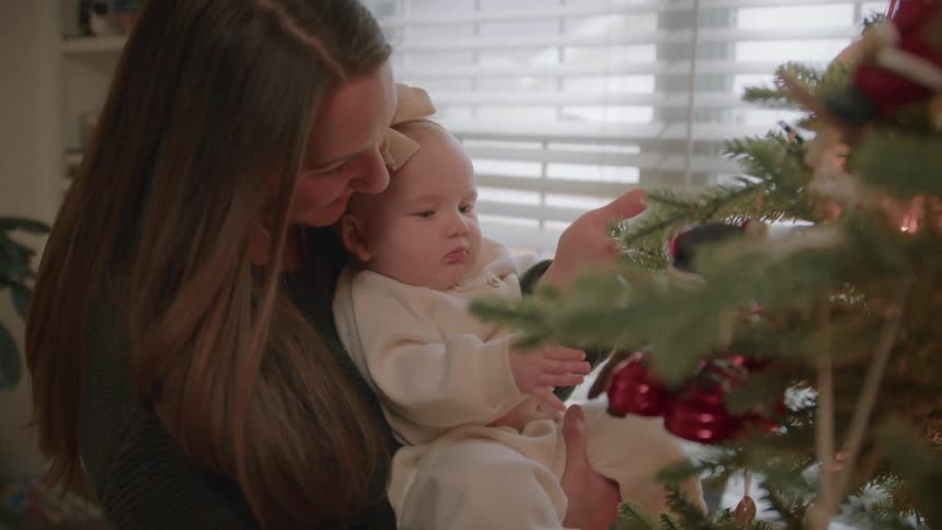 A loving mother holds her baby daughter and shows her the ornaments on their Christmas tree for the first time