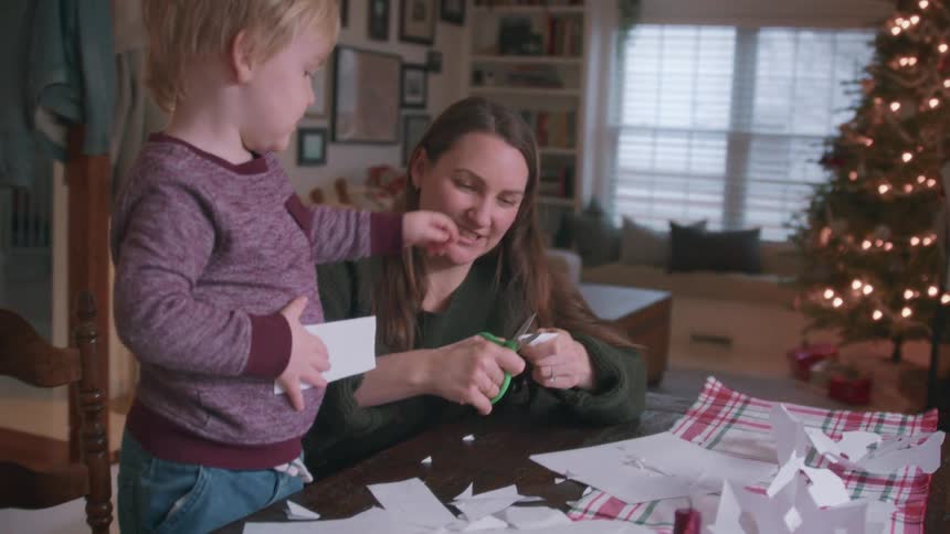 A mother and her toddler son make paper decorations together at a table in their cozy home, with a Christmas tree in the background
