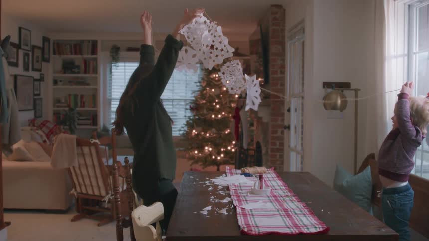 A loving mother and her adorable son stretch out a handmade paper snowflake garland in their home decorated for the holidays