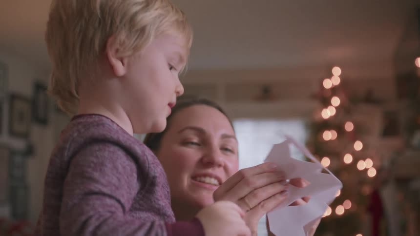 A loving mother and her toddler son share a heartwarming moment making a paper snowflake by the Christmas tree