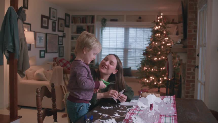 A mother and son laugh and smile while making paper snowflakes for Christmas, sharing a kiss in their cozy home with a tree in the background