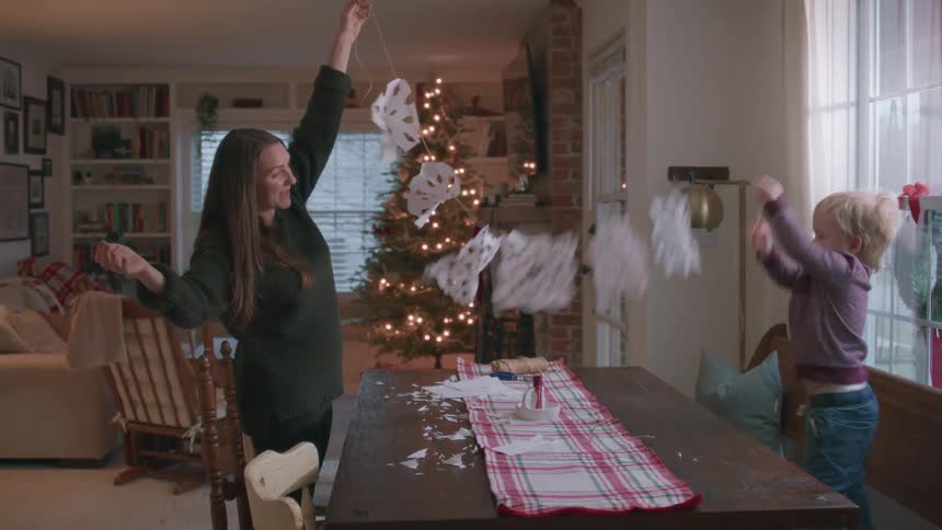 A joyful mother and her toddler son play with a handmade paper snowflake garland in their festively decorated home at Christmas time