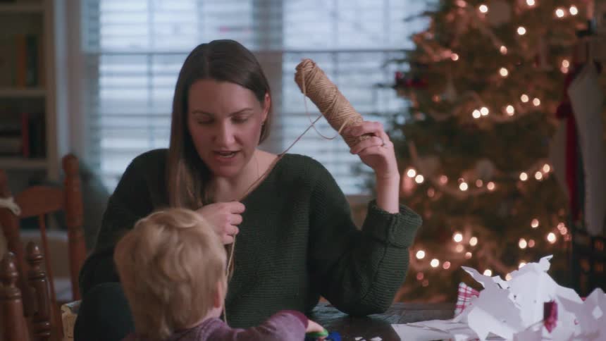 A mother and her young son share a sweet moment crafting holiday decorations with paper and twine in their cozy home, with a twinkling Christmas tree in the background