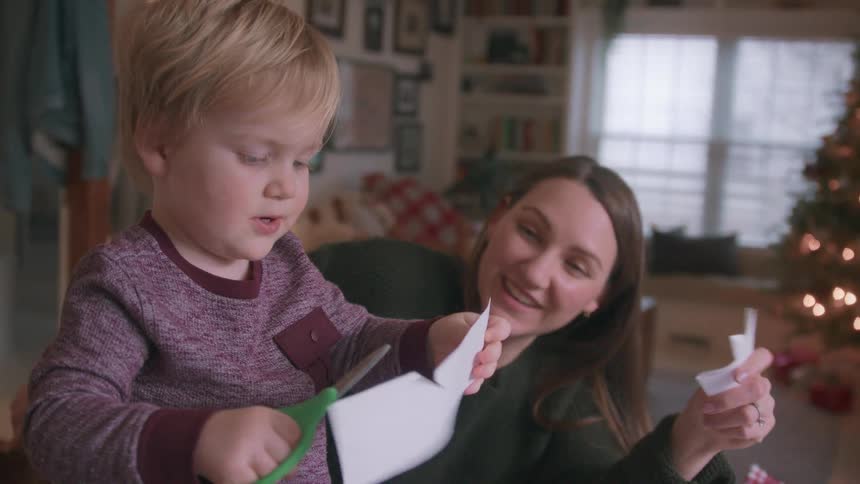 A mother helps her toddler son learn to use scissors while making paper crafts for Christmas in their warmly lit home