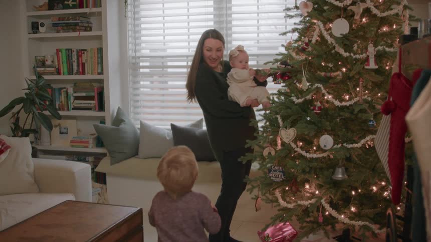 A mother holds her baby girl up to the decorated Christmas tree while her toddler son watches in their cozy living room