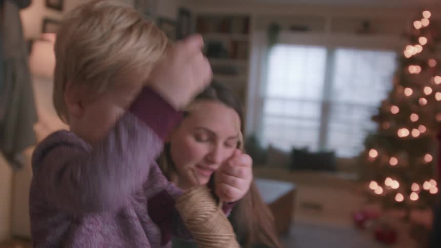A mother and her curious young son play with a spool of twine while making Christmas crafts together by a decorated tree
